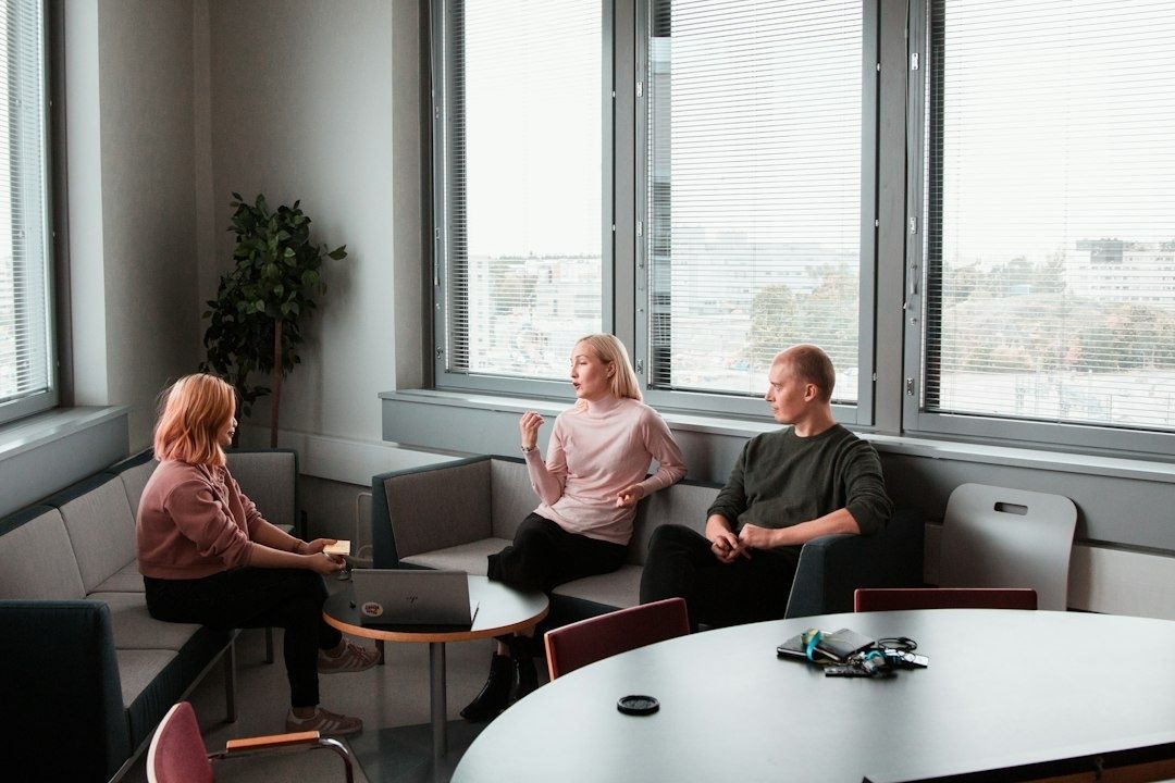 Tre personer sitter i samtal i ett ljust rum med stora fönster